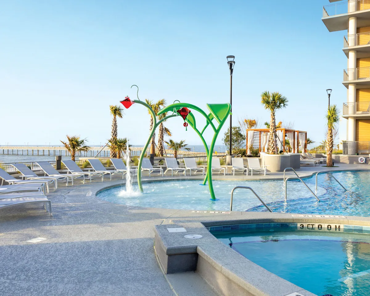 This vibrant exterior shot showcases a colorful water play structure surrounded by a spacious swimming pool, ample lounge chairs, palm trees, and an expansive ocean view in the background. The playful atmosphere is enhanced by the presence of children enjoying water play alongside adults relaxing poolside.