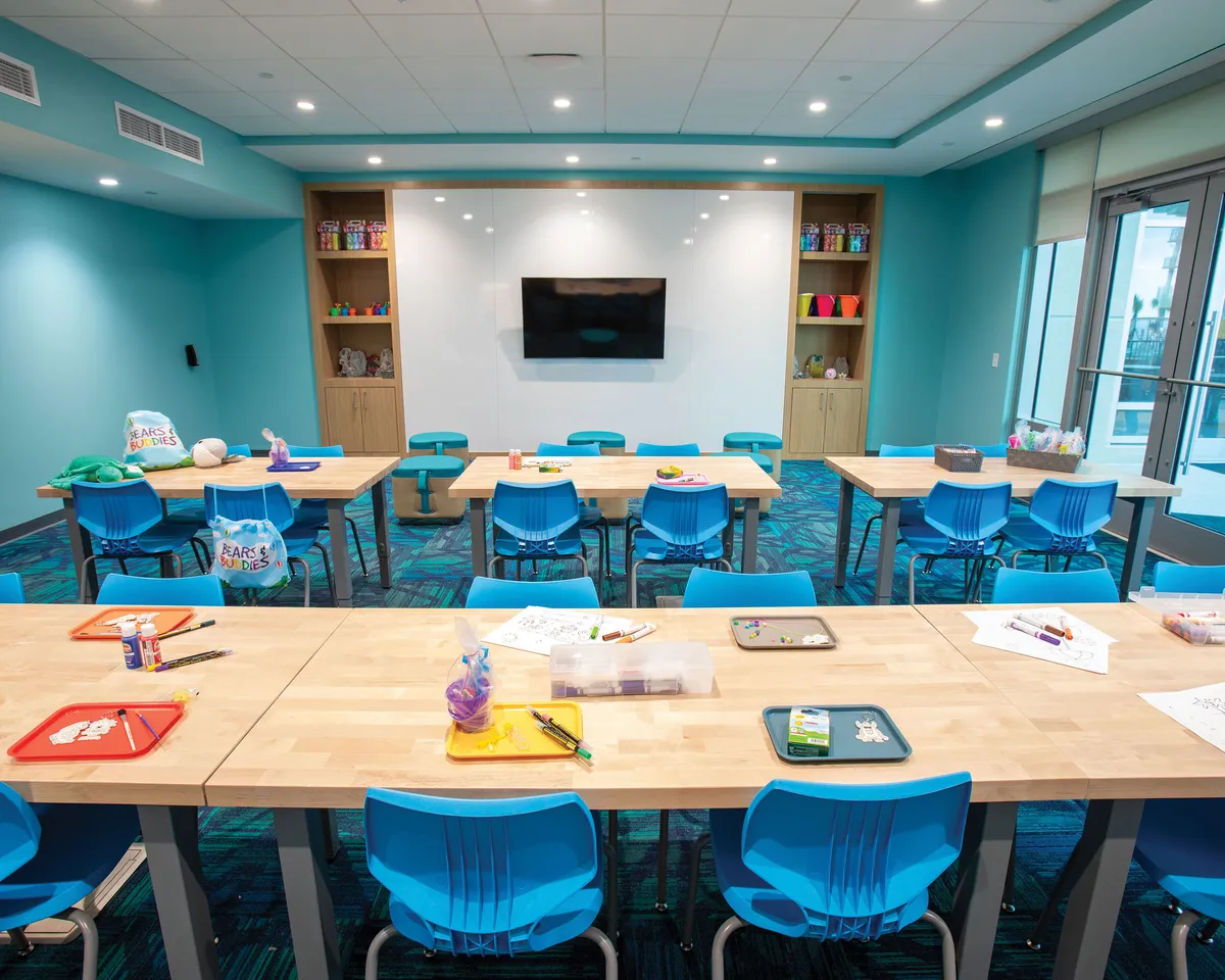 A playful, colorful room with tables set up for crafts, vibrant chairs, and shelves lined with a variety of items. While primarily a craft space, the communal tables and cheerful ambiance evoke the feel of shared meals or family gatherings around a table.