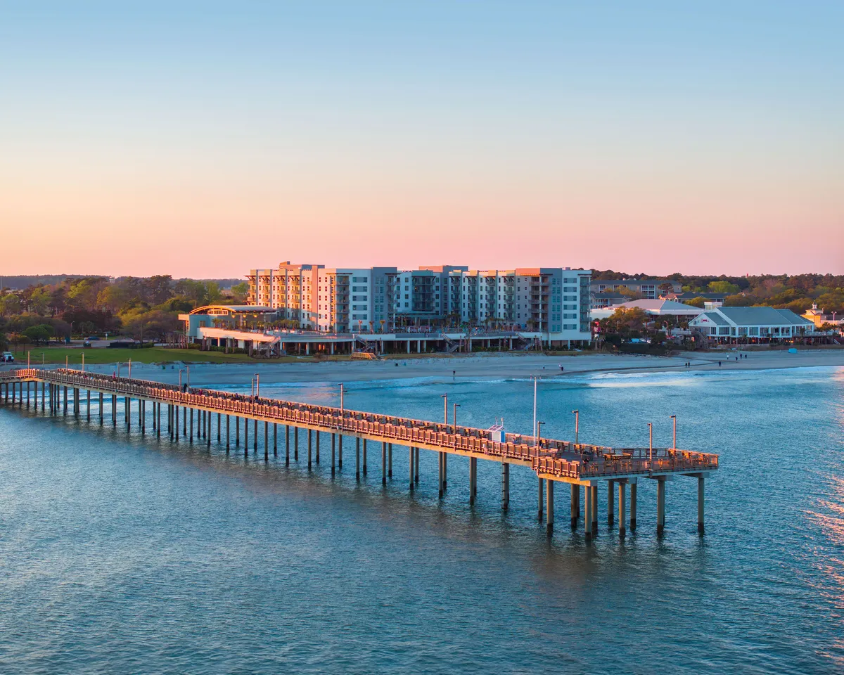 A charming Myrtle Beach pier lined with waterfront buildings under a glowing sunset sky. People stroll along the pier, some fishing while others dine at waterside restaurants, capturing the essence of evening adventures and local attractions.