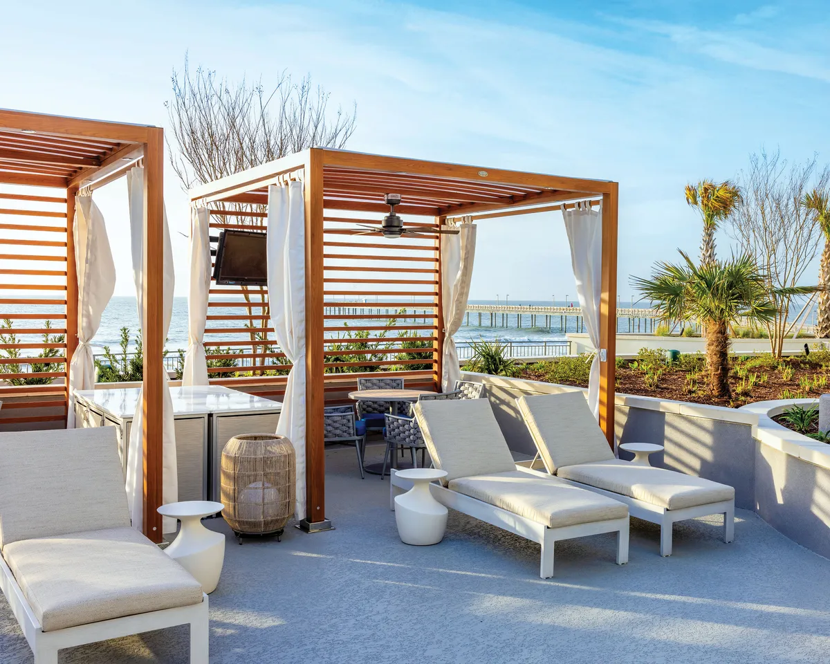 A tranquil poolside scene with elegant cabana structures, comfortable lounge chairs, and a serene water view fringed by palm trees. The setting is ideal for sunbathing, socializing, or sipping drinks in a relaxed, upscale environment.