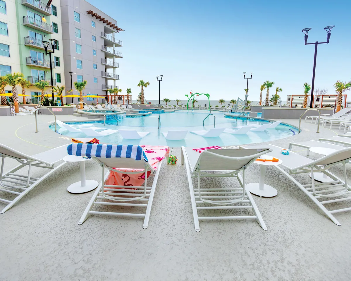 A vibrant, modern swimming pool area surrounded by poolside loungers draped with colorful towels, lush palm trees, and a contemporary building backdrop. The atmosphere is lively and inviting, with families and groups enjoying the warm sunshine, swimming, and gathering together.