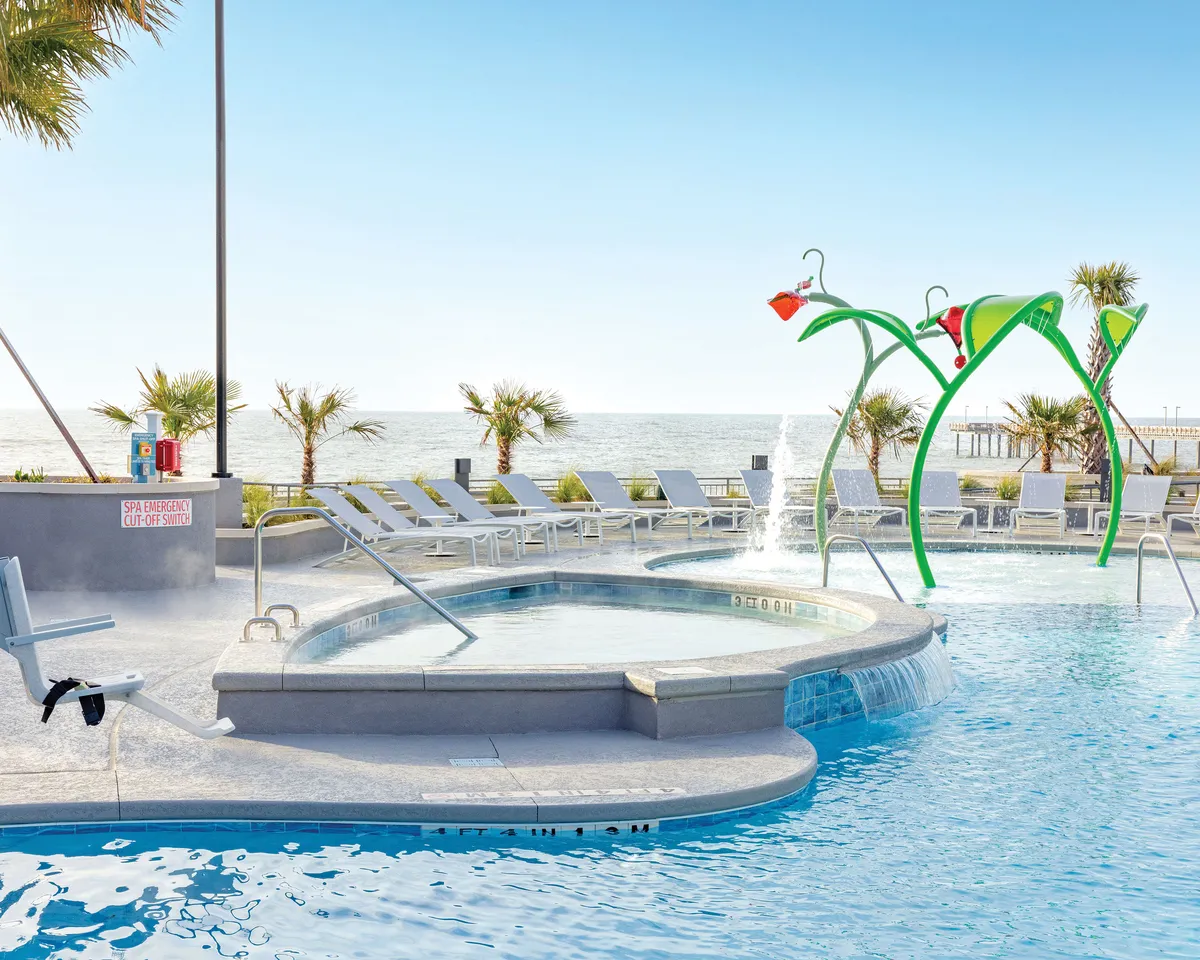 A spacious pool area featuring a dynamic water feature, plenty of comfortable lounge chairs, and sweeping views of the ocean framed by palm trees. Families are seen enjoying water play and gatherings, creating an atmosphere perfect for building vacation memories.