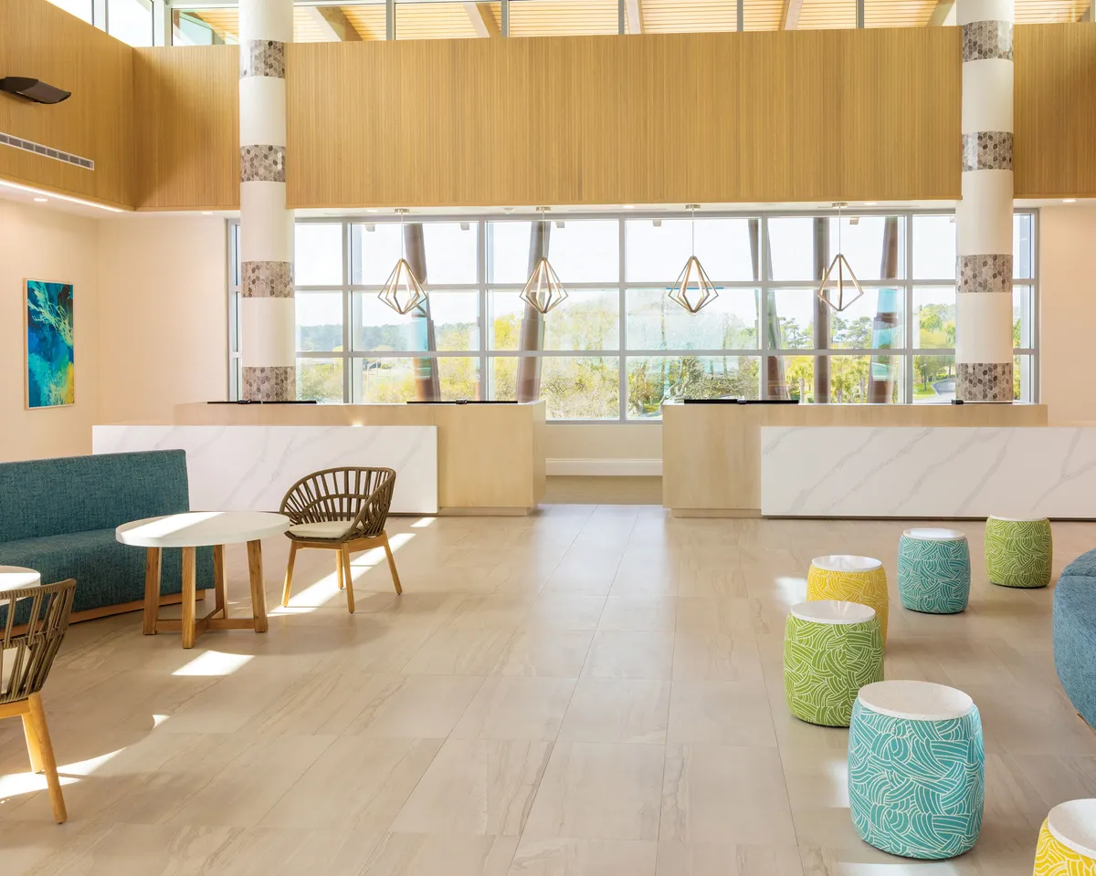 This image features a bright interior space bathed in natural light streaming through large windows. The room includes colorful, inviting seating options arranged for group gatherings, and a stylish reception desk adding a touch of coastal chic. The overall mood is welcoming, vibrant, and conducive to socializing, echoing the living area's function as a communal heart of the home.