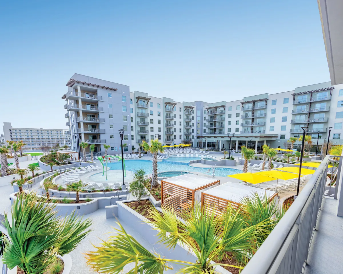 This image captures a vibrant resort pool area surrounded by palm trees and modern architecture, with inviting yellow umbrellas and guests lounging or swimming. The poolside bar is visible, offering refreshing drinks, while the sunset glow creates a relaxed, luxurious atmosphere ideal for socializing or unwinding after a day of exploring the beach.