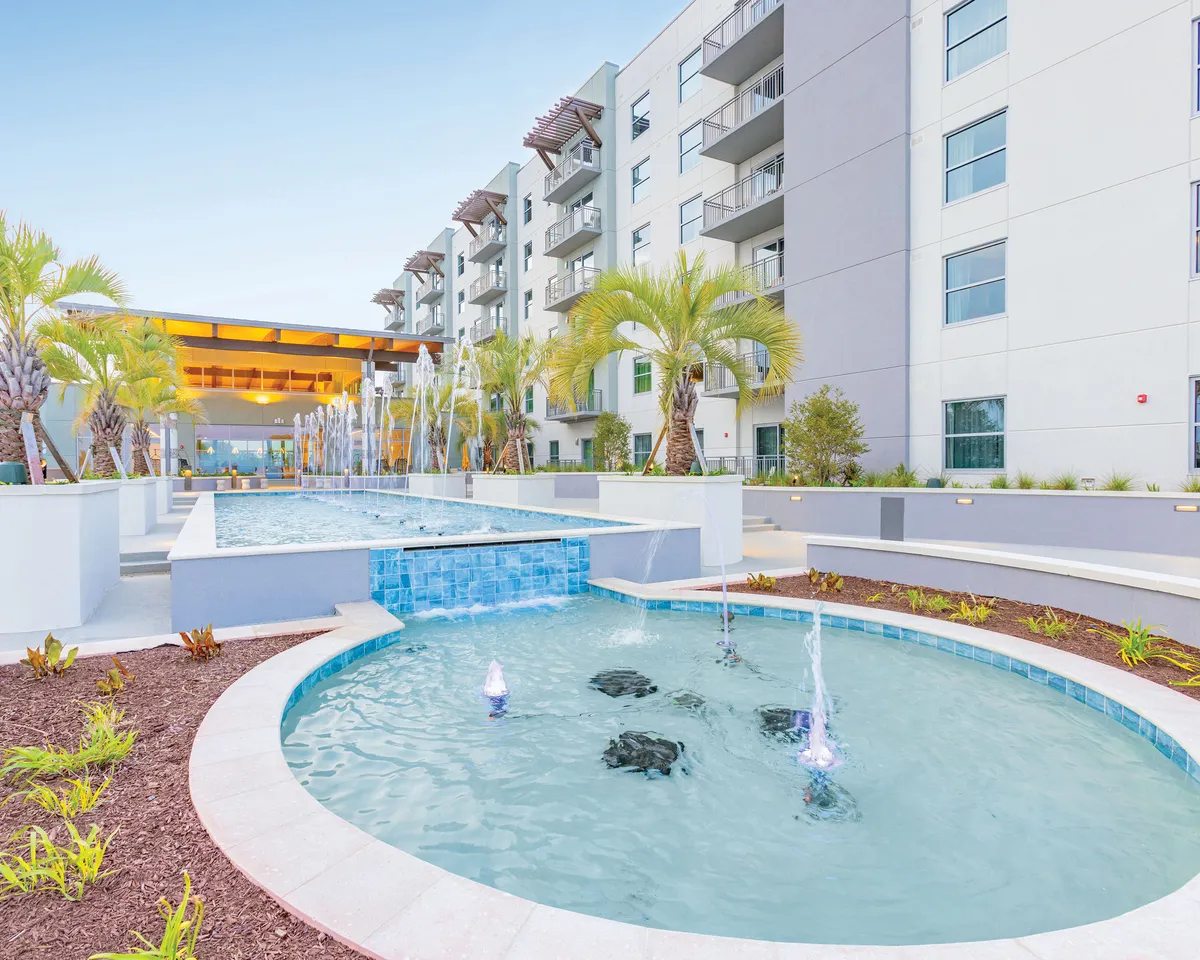 A vibrant outdoor scene featuring a sparkling swimming pool surrounded by water fountains, lush palm trees, and modern resort architecture. Groups of people are seen relaxing poolside and socializing in the open-air setting, with plenty of space for gatherings and outdoor fun.