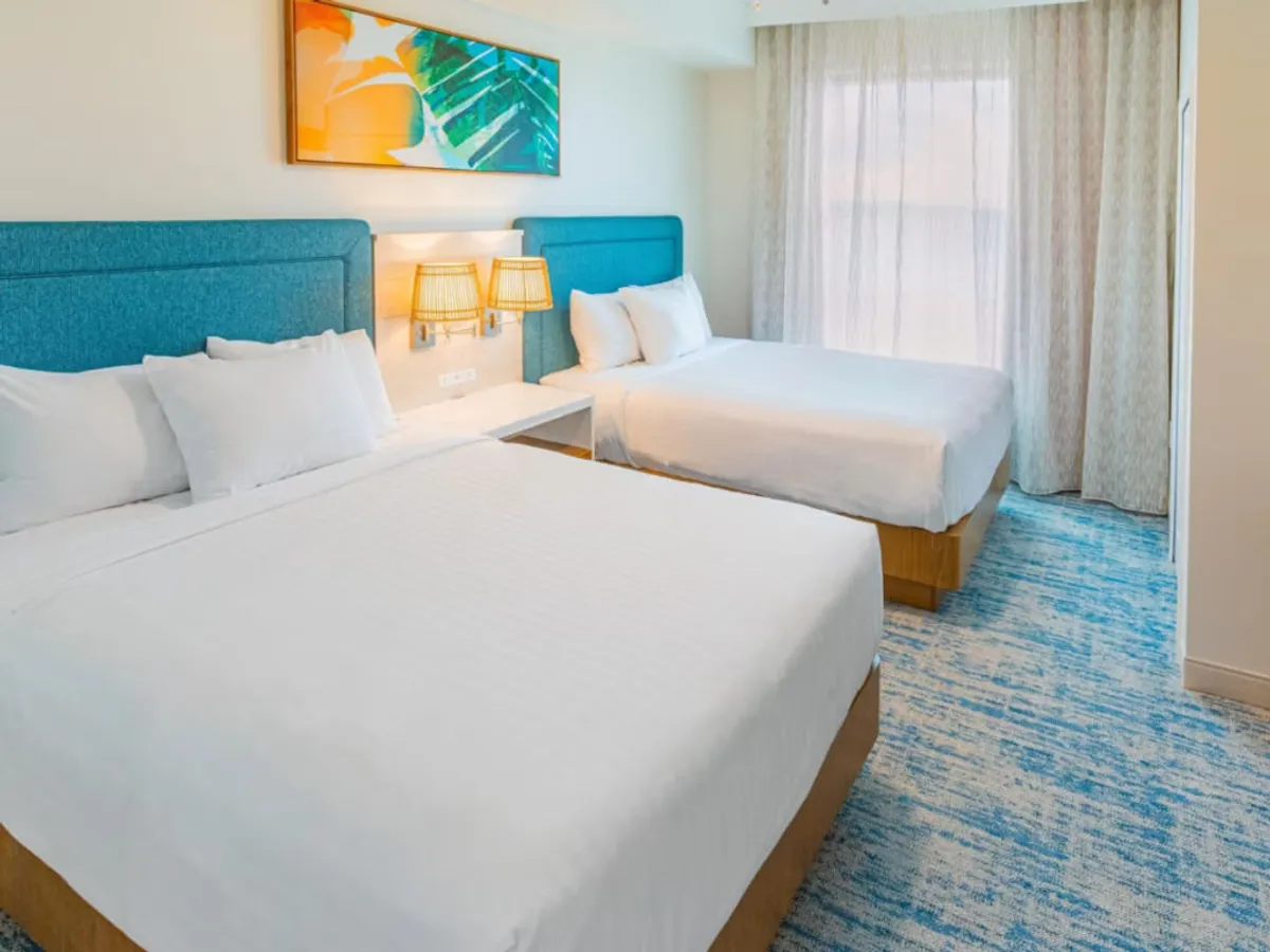 A spacious bedroom with two beds dressed in white linens, colorful wall art above the headboards, bedside lamps for evening reading, and a large window draped in sheer curtains that lets in plentiful natural light. The setup is cozy and ideal for kids, teens, or extra guests.